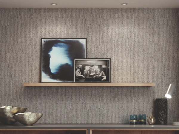 Modern living room wall with textured grey wallpaper, wooden floating shelf displaying two framed artworks including an abstract blue and white image and a black and white photograph, recessed ceiling lights, and wooden cabinetry below decorated with metallic bowls, decorative objects and a desk lamp.