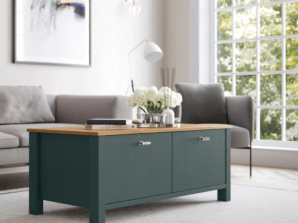 Modern living room with a dark teal coffee table featuring a wooden top and two drawers, styled with white roses in a glass vase, stacked books and decorative objects, positioned in front of a grey upholstered sofa, with a white arc floor lamp, framed artwork on the wall, and large windows overlooking green foliage.