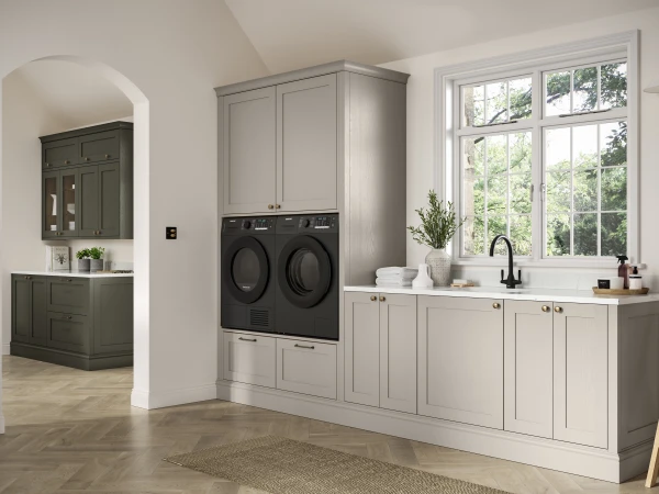 Contemporary laundry room with soft grey cabinetry, two black front-loading washing machines mounted above storage drawers, a white sink with black tap, large multi-pane window overlooking greenery, wooden ladder, potted plants, and an arched doorway leading to a dark green bathroom area beyond.