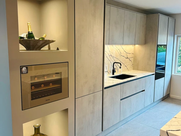 Modern kitchen with light wood cabinetry, marble backsplash and countertops, black sink with matte faucet, integrated appliances including built-in oven, and illuminated alcoves displaying champagne bottles and decorative items, with view to outdoor pool area through glass doors.